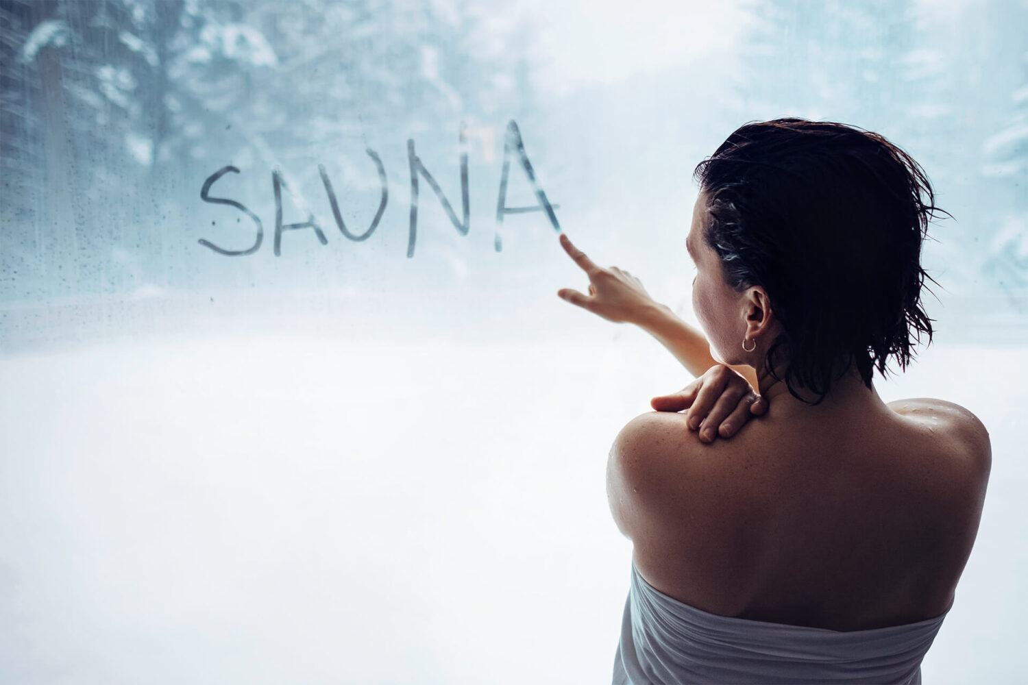Eine Frau schreibt mit ihrem Finger auf einem beschlagene Saunafenster das Wort "SAUNA".