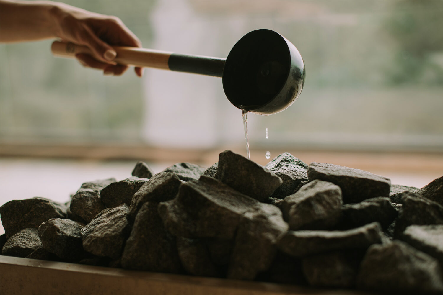 Frau gießt Wasser auf Steine in einem Ofen mit einer Schöpfkelle. Traditionelle Sauna.